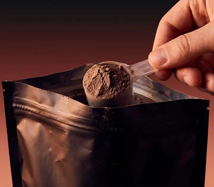 Hand lifting a scoop of chocolate superfood powder from a resealable foil pouch against a warm black-to-terracotta gradient