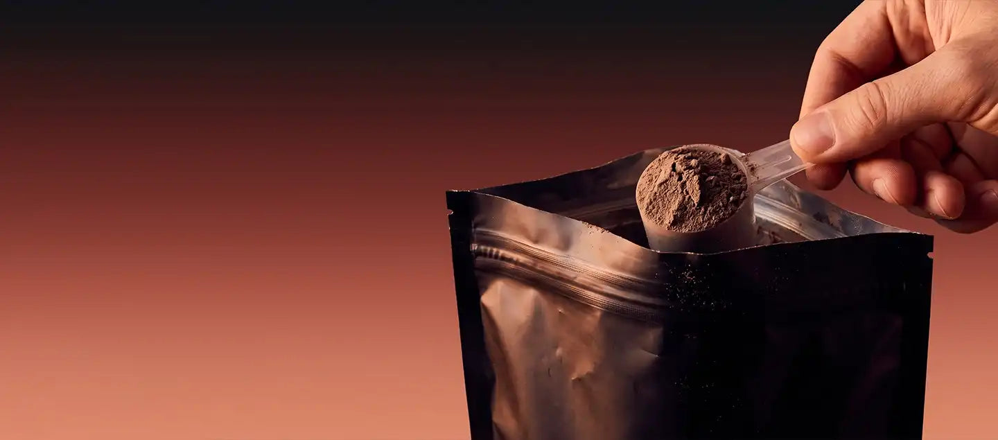 Hand lifting a scoop of chocolate superfood powder from a resealable foil pouch against a warm black-to-terracotta gradient