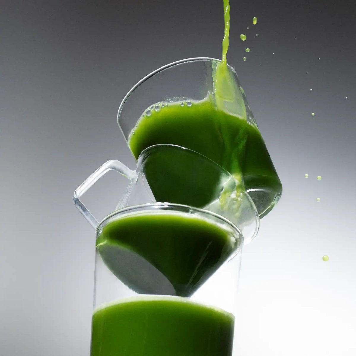 Organic ceremonial grade matcha pouring into a stack of clear glass mugs, with bright green droplets splashing against a grey gradient background