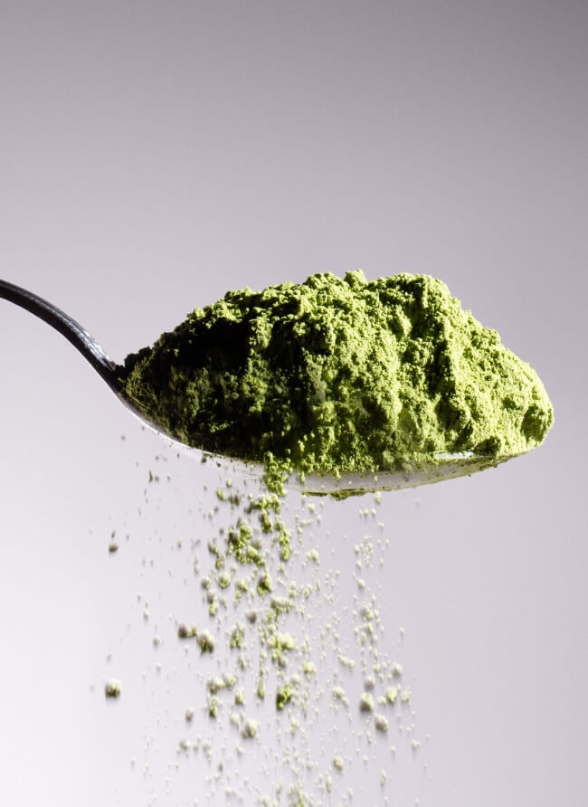 Heaping spoonful of organic green matcha powder with fine particles falling, set against a soft grey gradient background.