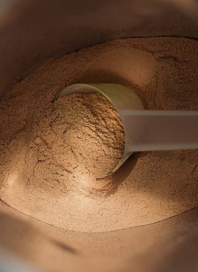 Overhead close-up of a scoop resting in a large batch of light brown superfood powder, lit by warm, soft light