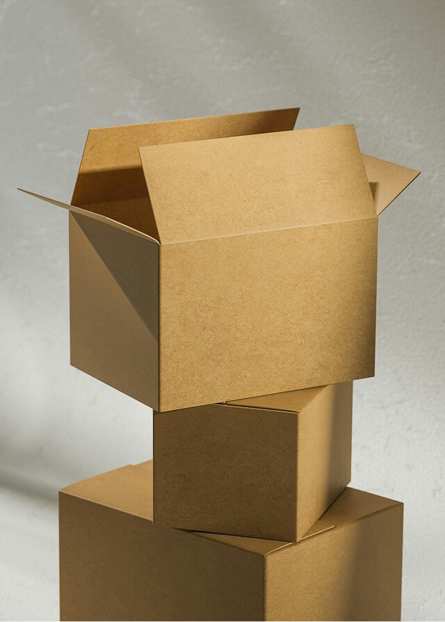 Three stacked kraft cardboard shipping boxes, the top one open, in soft sunlight against a textured light grey wall